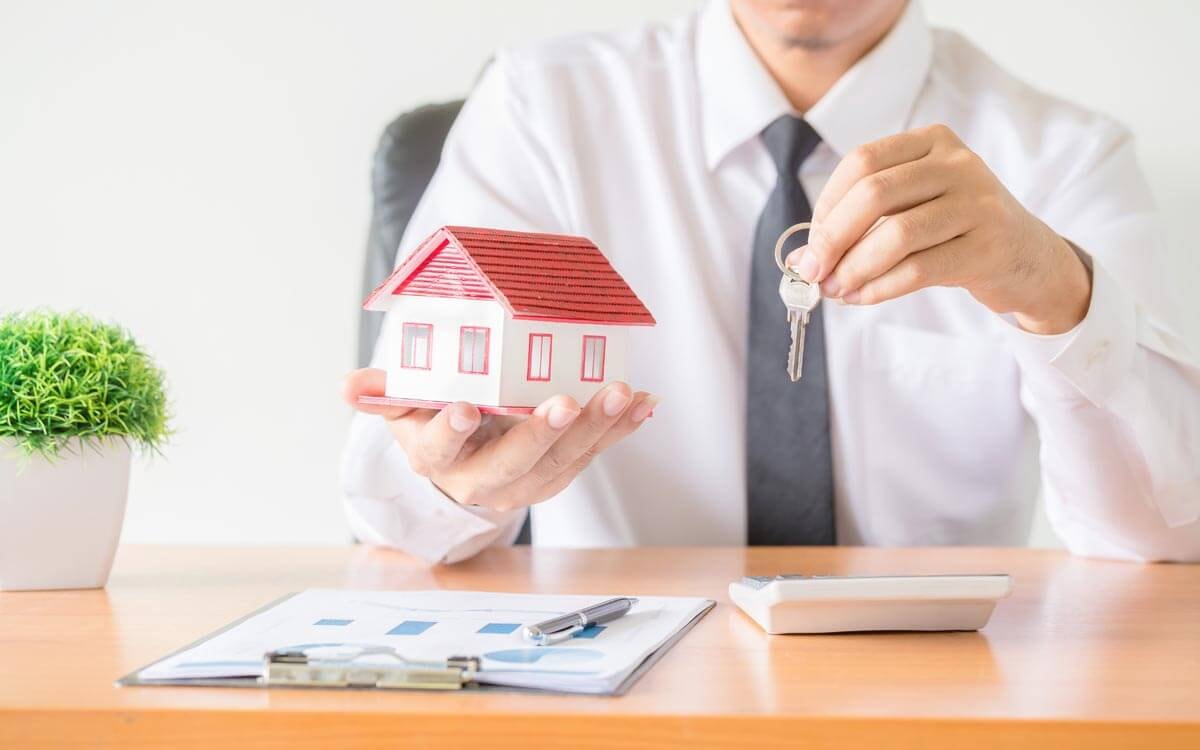 Nahaufnahme eines Immobilienmaklers an einem Schreibtisch, der in der einen Hand die Miniatur eines Hauses mit rotem Satteldach und in der anderen Hand einen Haustürschlüssel hoch hält.